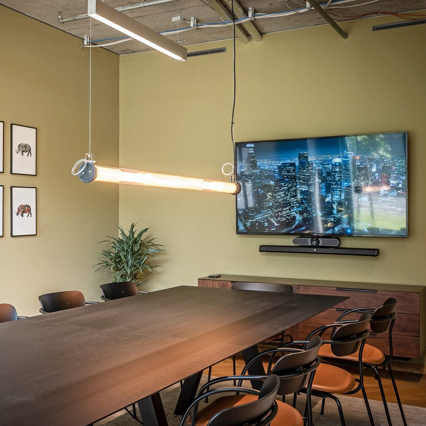 Ein moderner Konferenzraum mit einem langen, dunklen Holztisch, umgeben von schwarzen Stühlen. Die Wände sind olivgrün und an einer Seite hängt dekorative Kunst. Ein großer Fernseher zeigt eine Stadtlandschaft. Darüber, ein Bild von EDGE Workspaces.