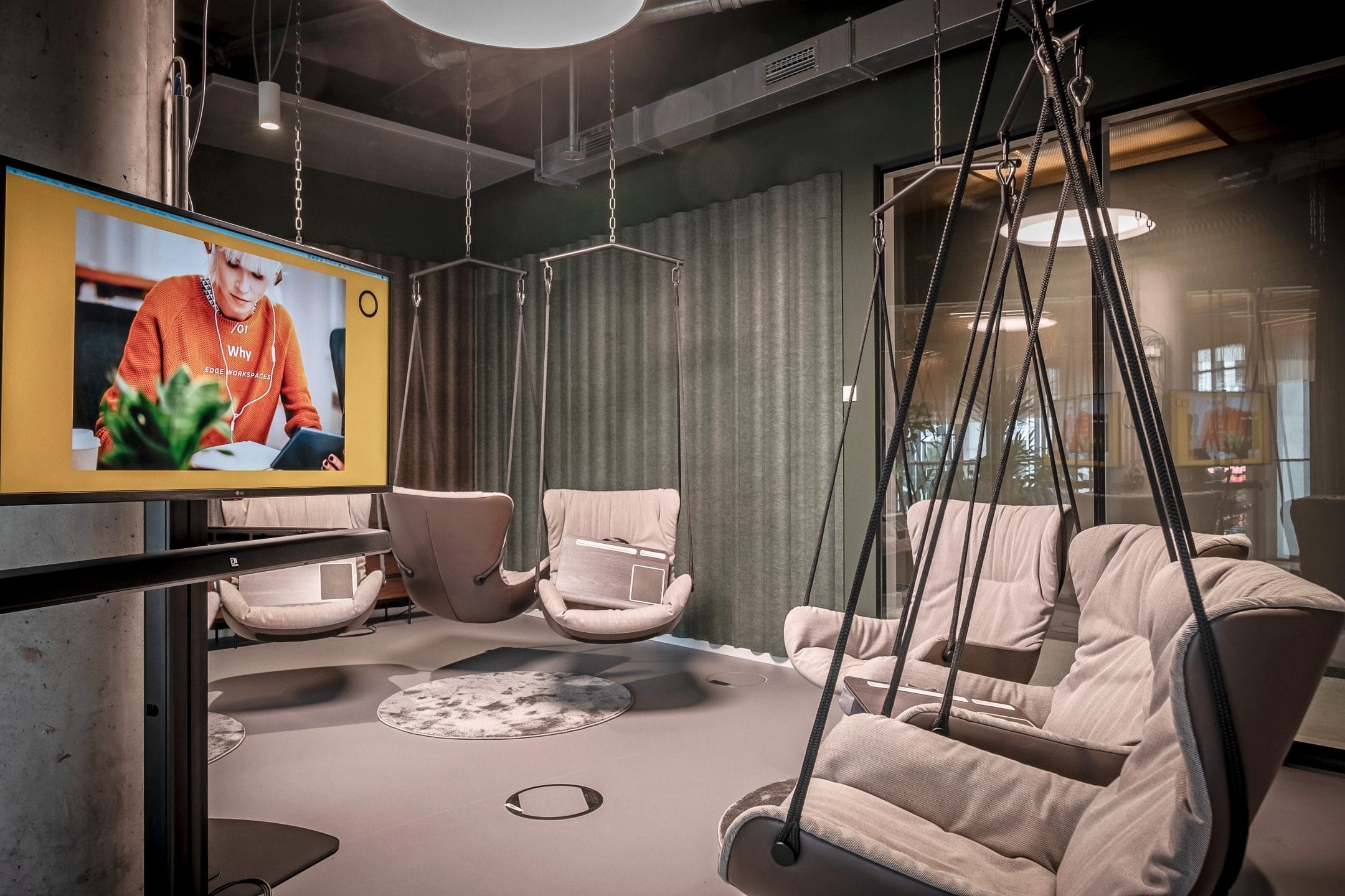 A modern office lounge featuring four suspended egg chairs with plush cushions, centered around a low circular table. The space is decorated with muted tones and an illuminated digital screen displays an image of a senior woman cooking An image from EDGE Workspaces.