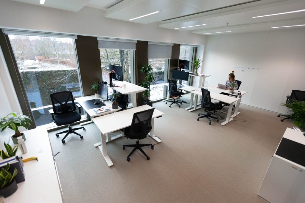 Ein geräumiges, gut beleuchtetes, modernes Büro mit großen Fenstern, mehreren Arbeitsplätzen mit Computern und einigen Mitarbeitern. Zur Ausstattung gehören Grünpflanzen und ergonomische Stühle. Ein Bild von EDGE Workspaces.