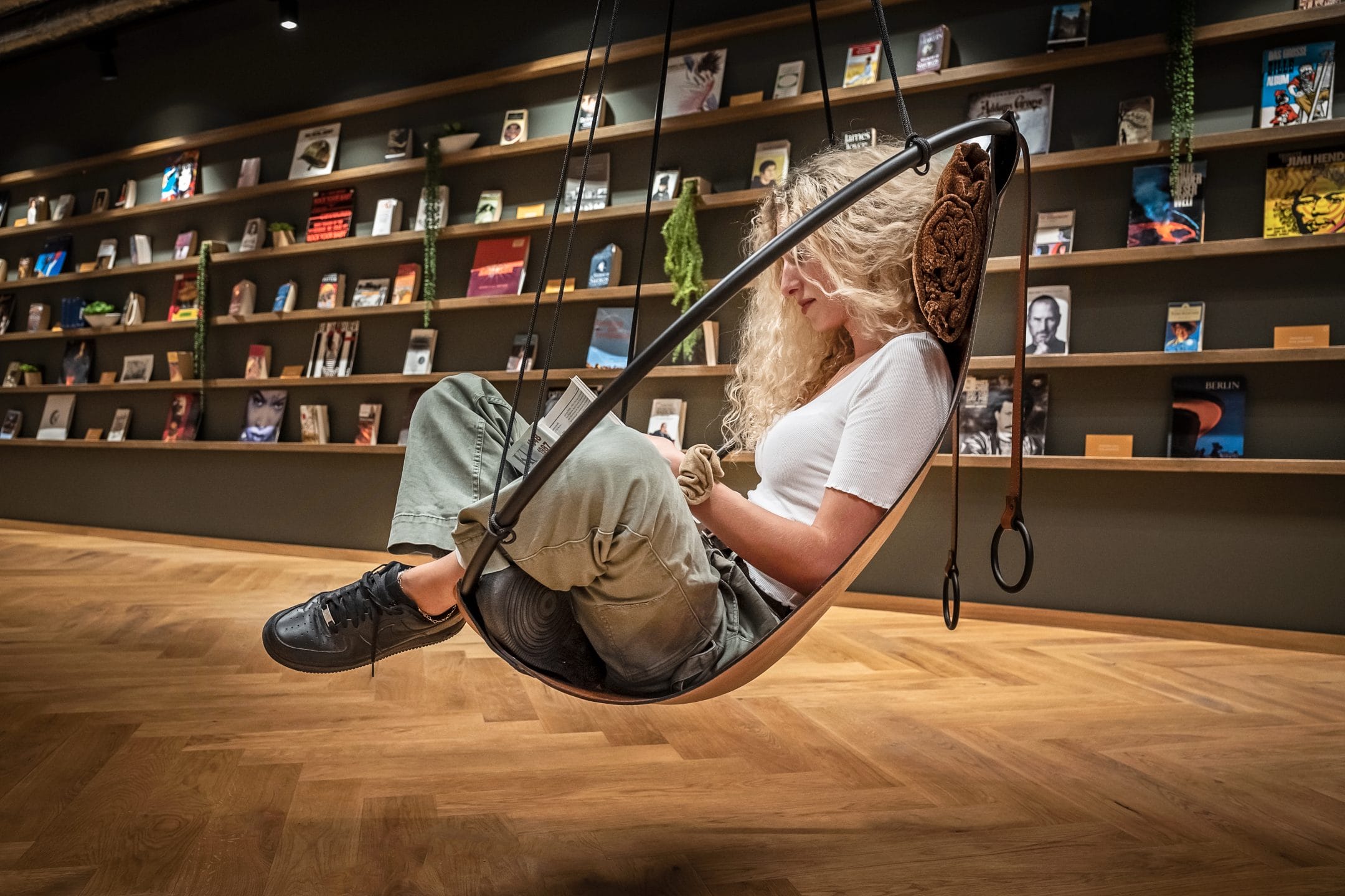 Eine Frau sitzt in einem stilvollen Schaukelstuhl und liest ein Buch in einer modernen Buchhandlung, die gleichzeitig als innovative Coworking-Umgebung dient, wobei die Wände von Regalen gesäumt sind, die mit Büchern gefüllt sind. Ihre entspannte Haltung und Konzentration deuten auf ein Bild von EDGE Workspaces hin.