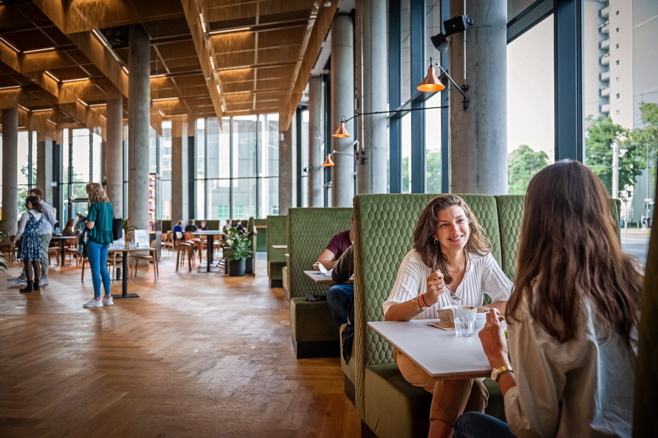 Two women are engaging in conversation at a table in a spacious, modern cafe with large windows and wooden beams, doubling as a coworking space. Other patrons chat or work in the background under warm lighting An image from EDGE Workspaces.