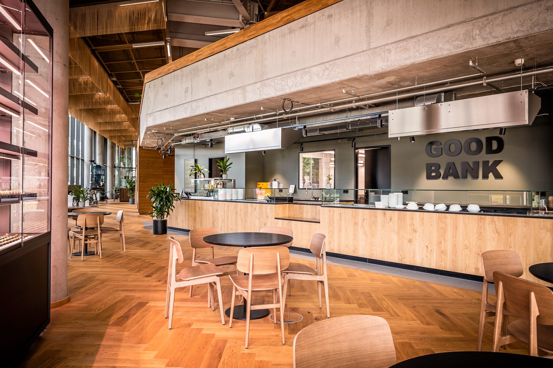 A modern and spacious interior of a restaurant named "Good Bank," featuring wooden tables, chairs, and a bench with overhead industrial lighting. A well-equipped kitchen bar with plants occupies the background, transitioning seamlessly An image from EDGE Workspaces.