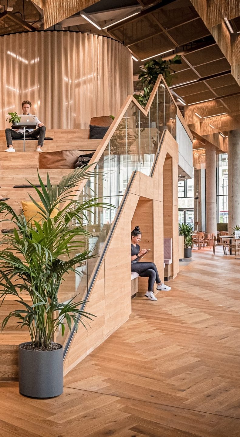 Ein modernes Bürointerieur mit einer Holztreppe mit integrierten Sitzgelegenheiten und speziellen Arbeitsbereichen. Einzelpersonen nutzen Laptops auf verschiedenen Ebenen, umgeben von Pflanzen und einem stilvollen, luftigen Design mit natürlichem Licht. Ein Bild von EDGE Workspaces.