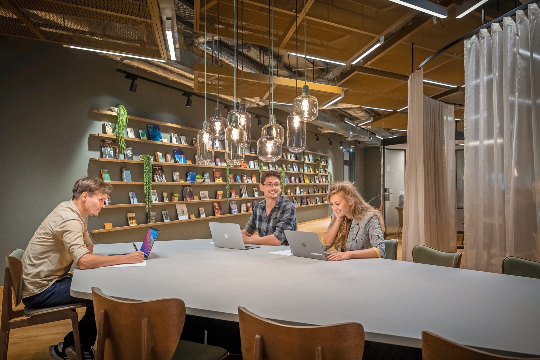 Drei Personen, zwei Männer und eine Frau, sitzen an einem langen Tisch in einem modernen Coworking Space und arbeiten an Laptops und einem Tablet. Regale mit Büchern und warm hängende Lampen darüber unterstreichen ein Bild von EDGE Workspaces.