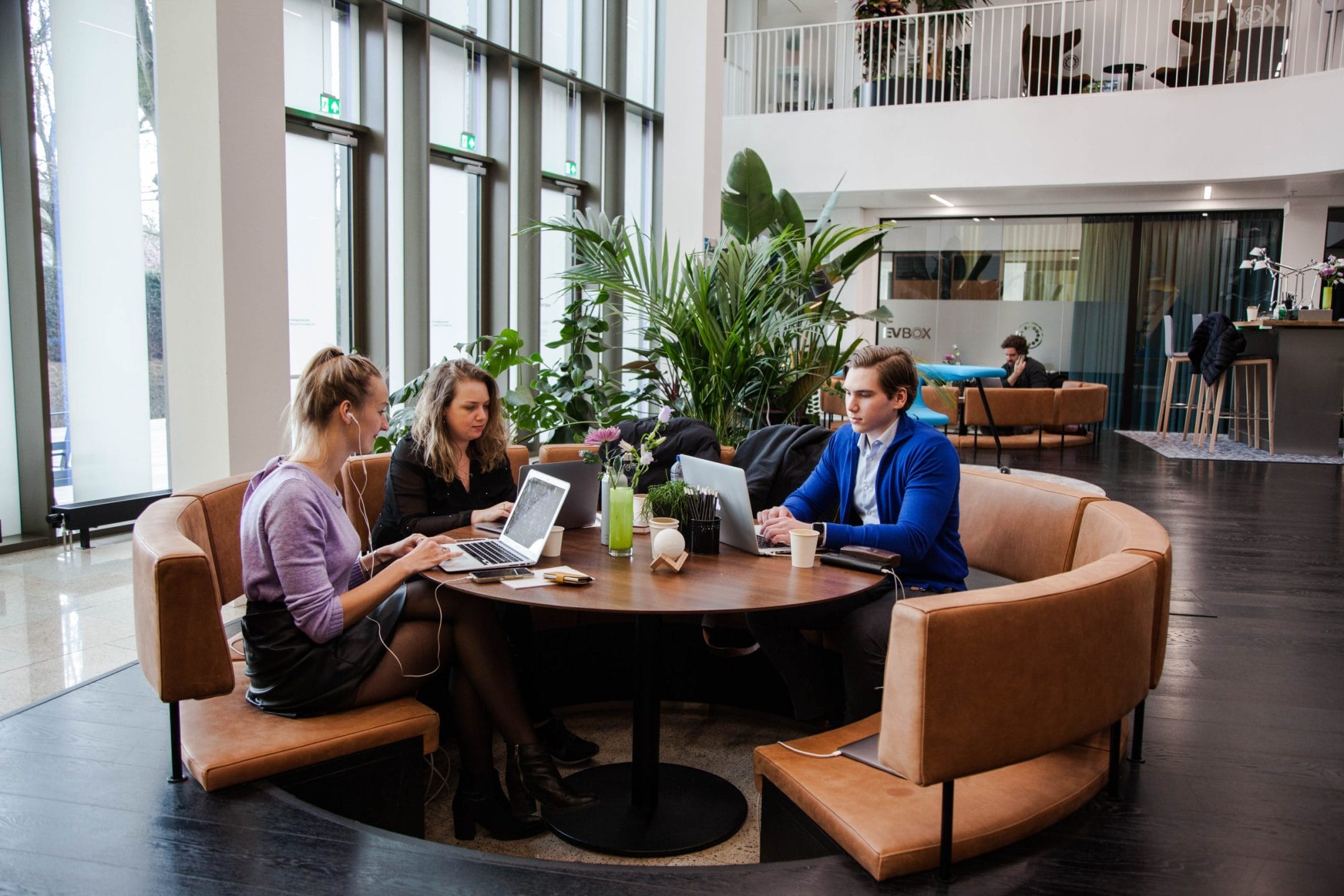 Three professionals, two women and one man, sit around a circular table in an innovative coworking environment, working on laptops and discussing. Large windows and green plants are visible in the background. An image from EDGE Workspaces.