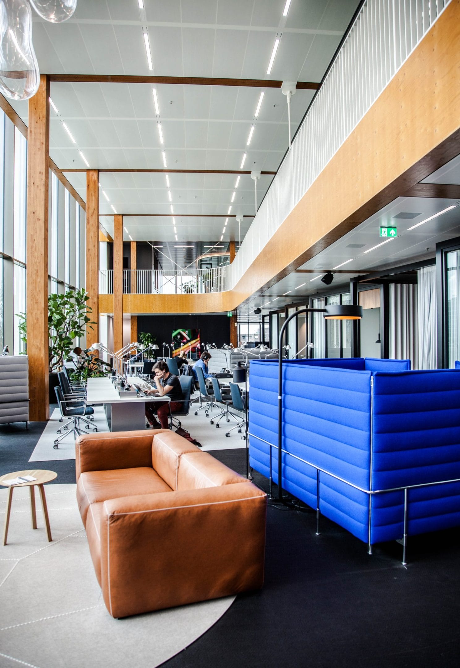 Modern office lobby with high ceilings, wooden panels, and natural light. It features stylish seating areas with brown leather sofas and blue padded booths, patterned rugs, and a coworking space with desks and An image from EDGE Workspaces.
