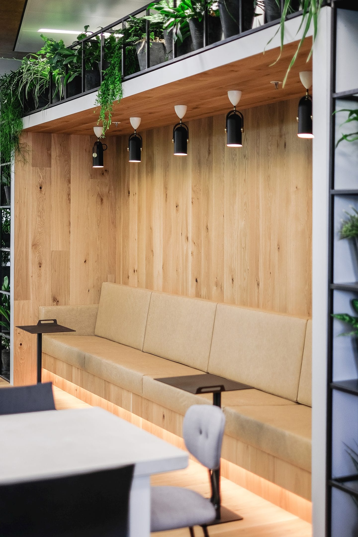 A cozy modern café corner with a continuous beige upholstered bench under a wooden alcove lined with hanging plants, transformed into an innovative coworking environment. Overhead, black pendant lights illuminate the area. An image from EDGE Workspaces.