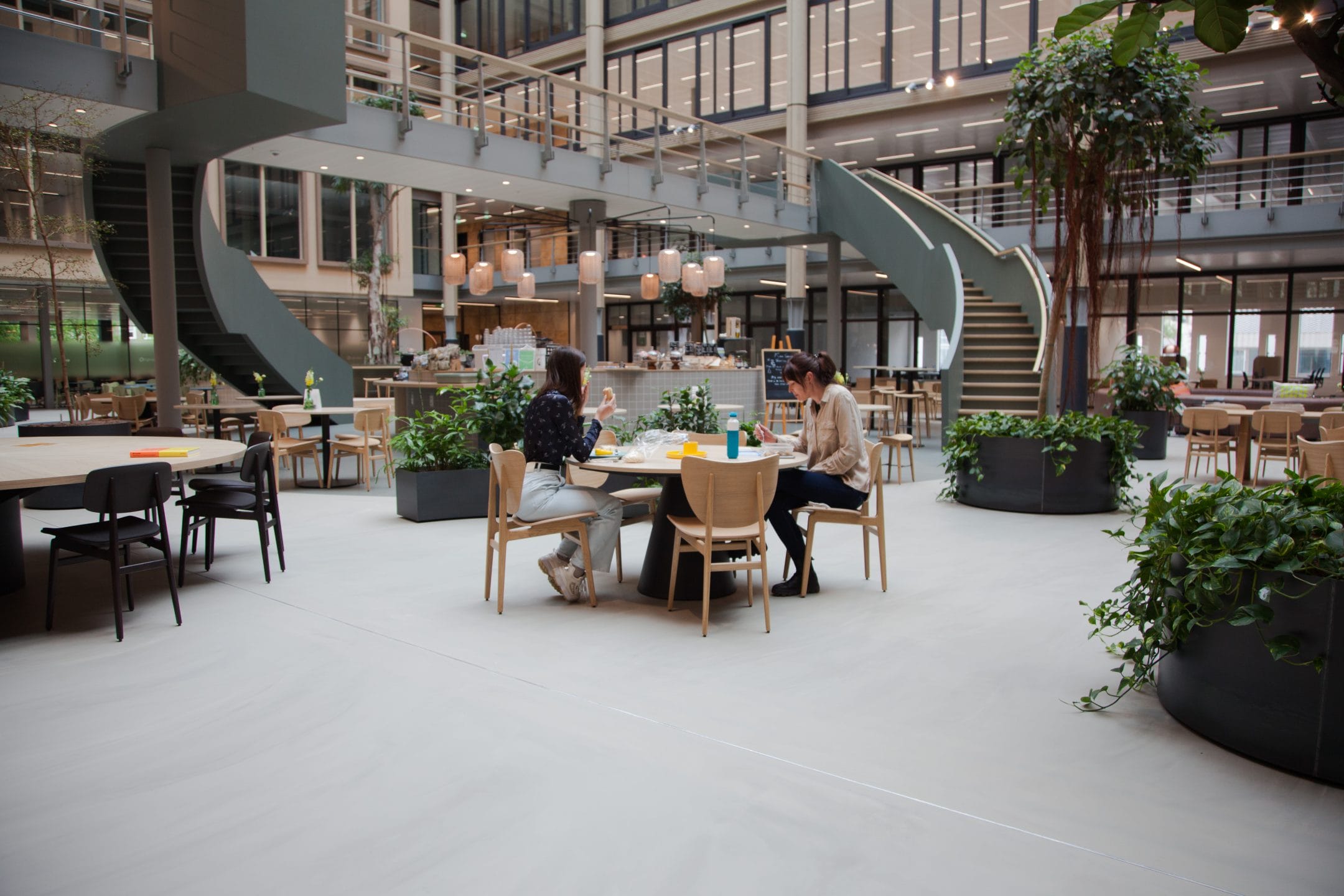 Spacious, modern cafeteria within an innovative coworking environment, featuring large green plants, varied seating arrangements, and two curved staircases. People are dining and conversing at tables.
 An image from EDGE Workspaces.