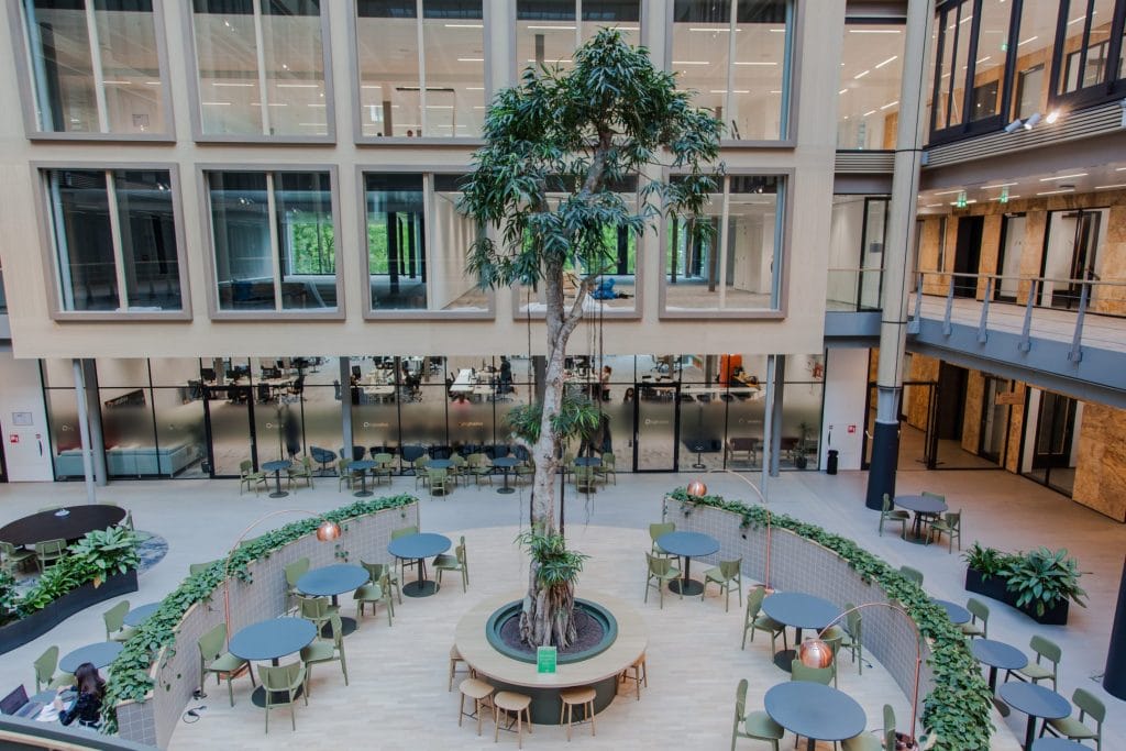 Innenansicht eines modernen Büroatriums mit großen Topfbäumen, umgeben von runden Sitzbereichen. Die oberen Etagen sind mit Glasfenstern und Balkonen ausgestattet. Der Raum ist als flexible Arbeitsplatzlösung mit einem Ein Bild von EDGE Workspaces.