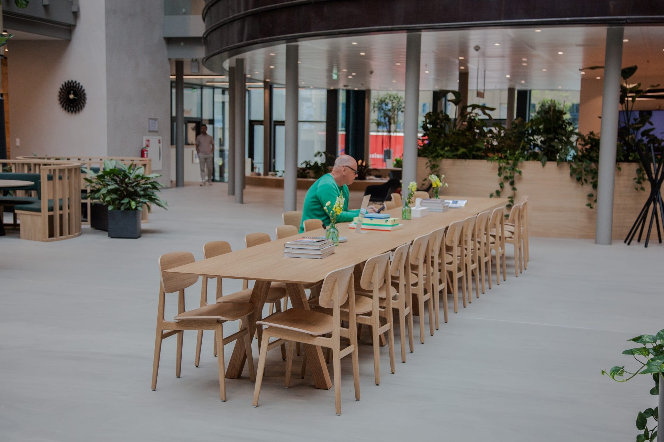 Ein geräumiges, modernes Büro-Atrium mit einem langen Holztisch mit passenden Stühlen, ideal für Coworking-Spaces. Ein älterer Mann in einem grünen Pullover liest am Tisch, umgeben von Büchern und Papieren. Ein Bild von EDGE Workspaces.