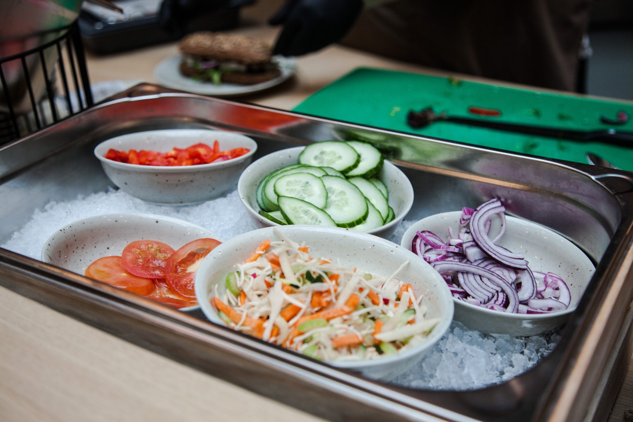 Ein Metalltablett mit kleinen Schalen mit frischen Gemüse-Toppings, darunter geschnittene Tomaten, Gurken, rote Zwiebeln und gemischter Salat auf einem Bett aus zerstoßenem Eis, mit einem teilweise sichtbaren Veggie-Burger im Bild. Ein Bild von EDGE Workspaces.