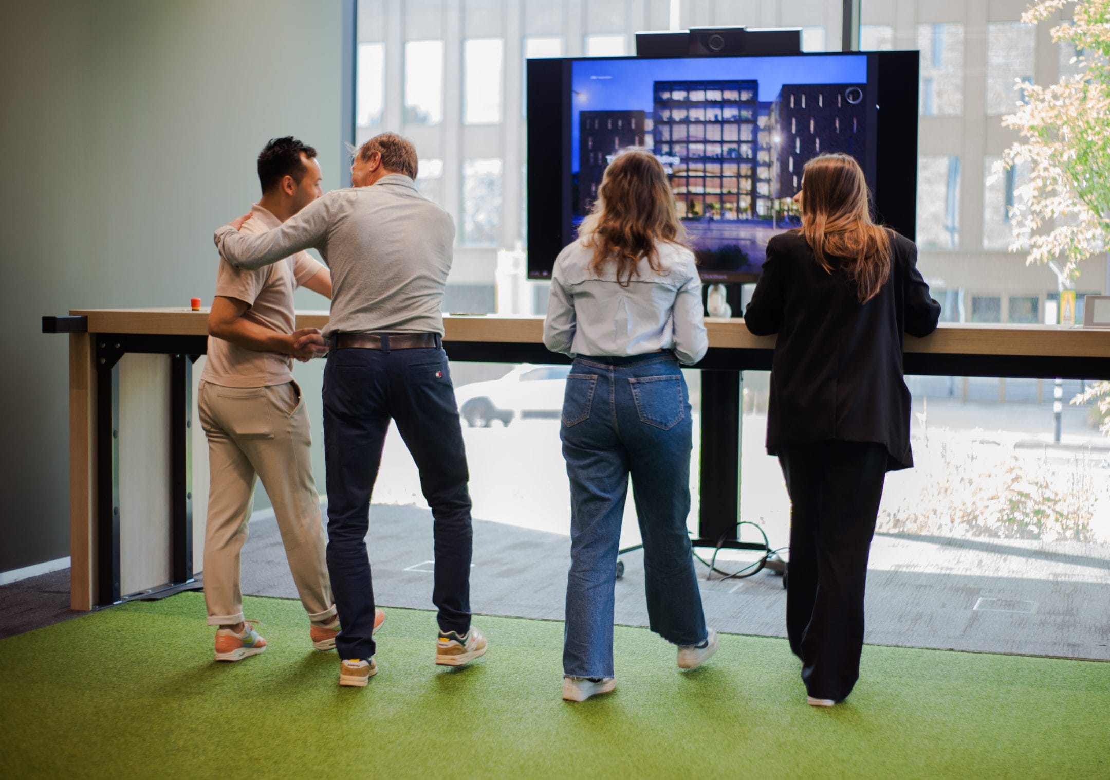 Vier Personen stehen zusammen in einem modernen Büro und betrachten einen großen Bildschirm, der ein Gebäudeabbild zeigt. Sie sind in eine Präsentation oder Diskussion vertieft, wobei der Fokus auf dem Bildschirm liegt, in einer innovativen Coworking-Umgebung. Ein Bild von EDGE Workspaces.