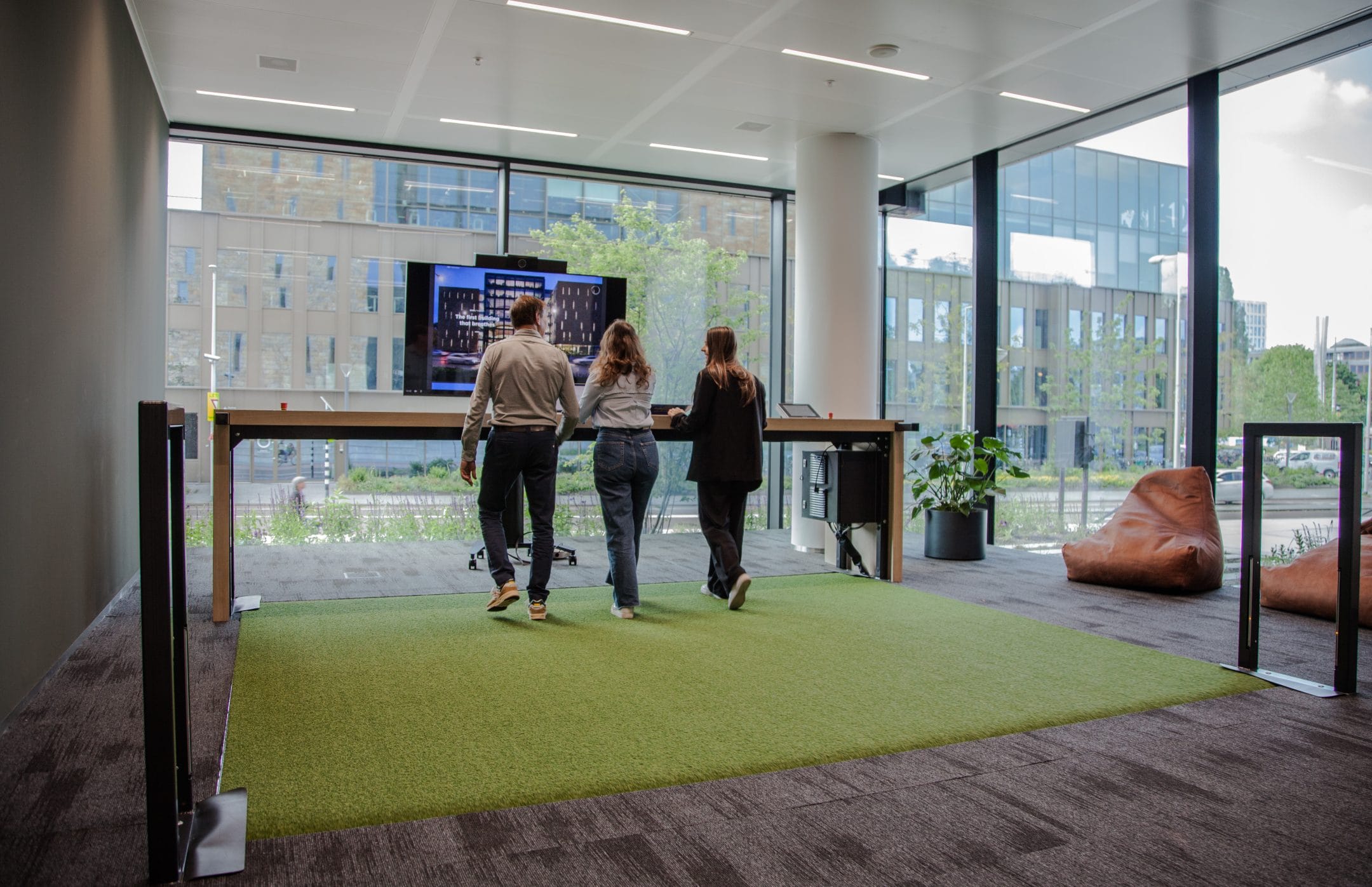 Three people stand at a digital display within a brightly lit sustainable office solutions with large windows, a green carpet, and relaxed seating nearby. The outside shows urban buildings and vegetation. An image from EDGE Workspaces.