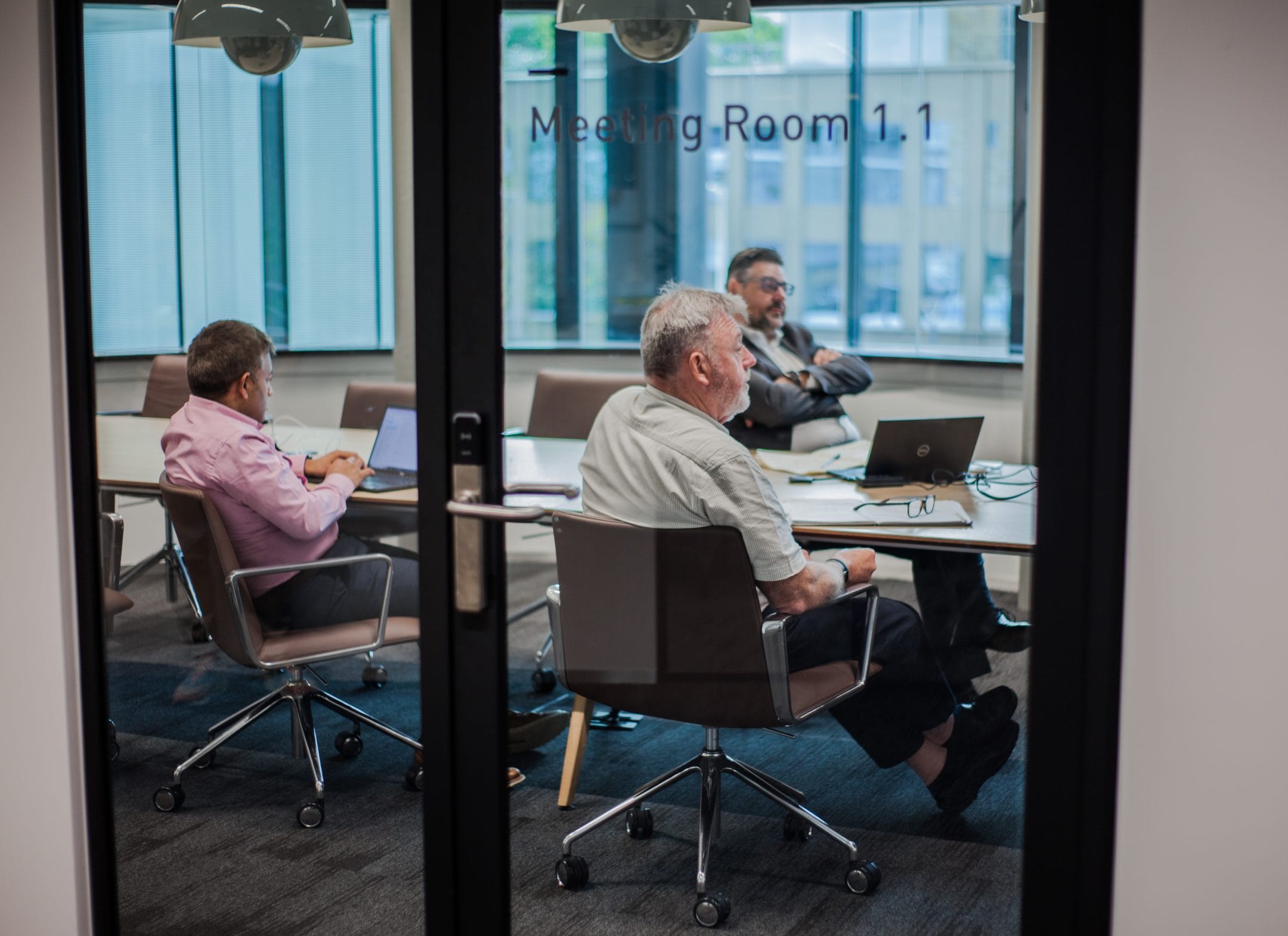 Drei Männer in einem modernen Besprechungsraum, gesehen durch eine Glastür mit der Aufschrift "Besprechungsraum 1.1" in einer innovativen Coworking-Umgebung. Zwei sitzen an einem Tisch vor einem Laptop, und ein Bild von EDGE Workspaces.