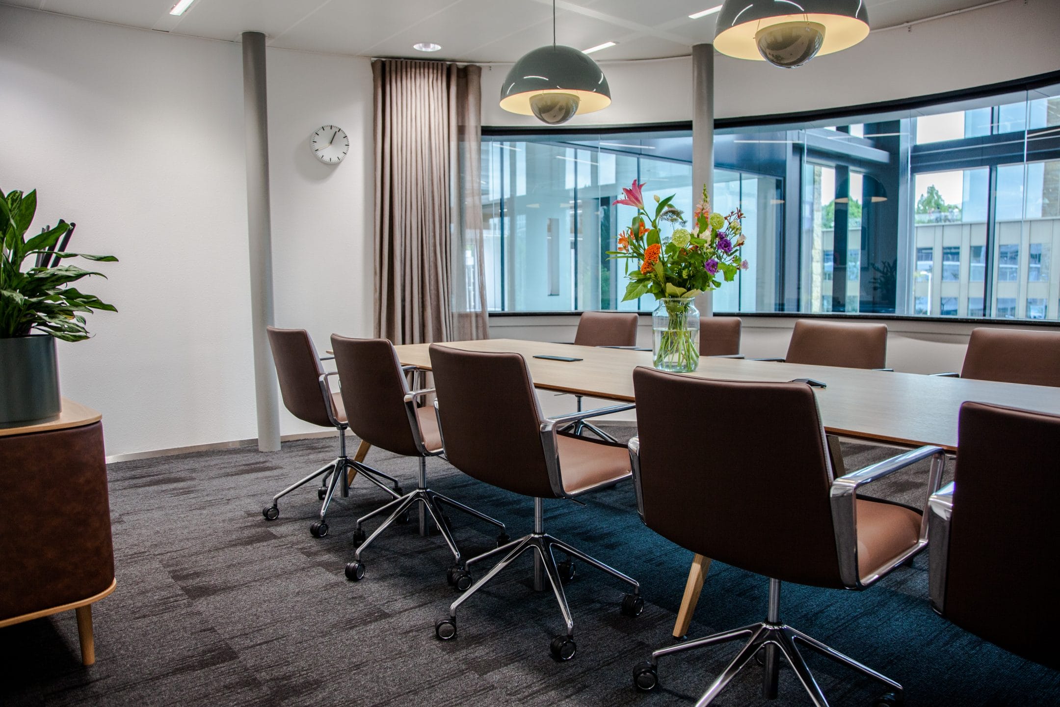 A modern office meeting room with a large wooden table surrounded by brown chairs. The room features contemporary lighting fixtures, potted plants, a large clock on the wall, and a window with blinds. This An image from EDGE Workspaces.