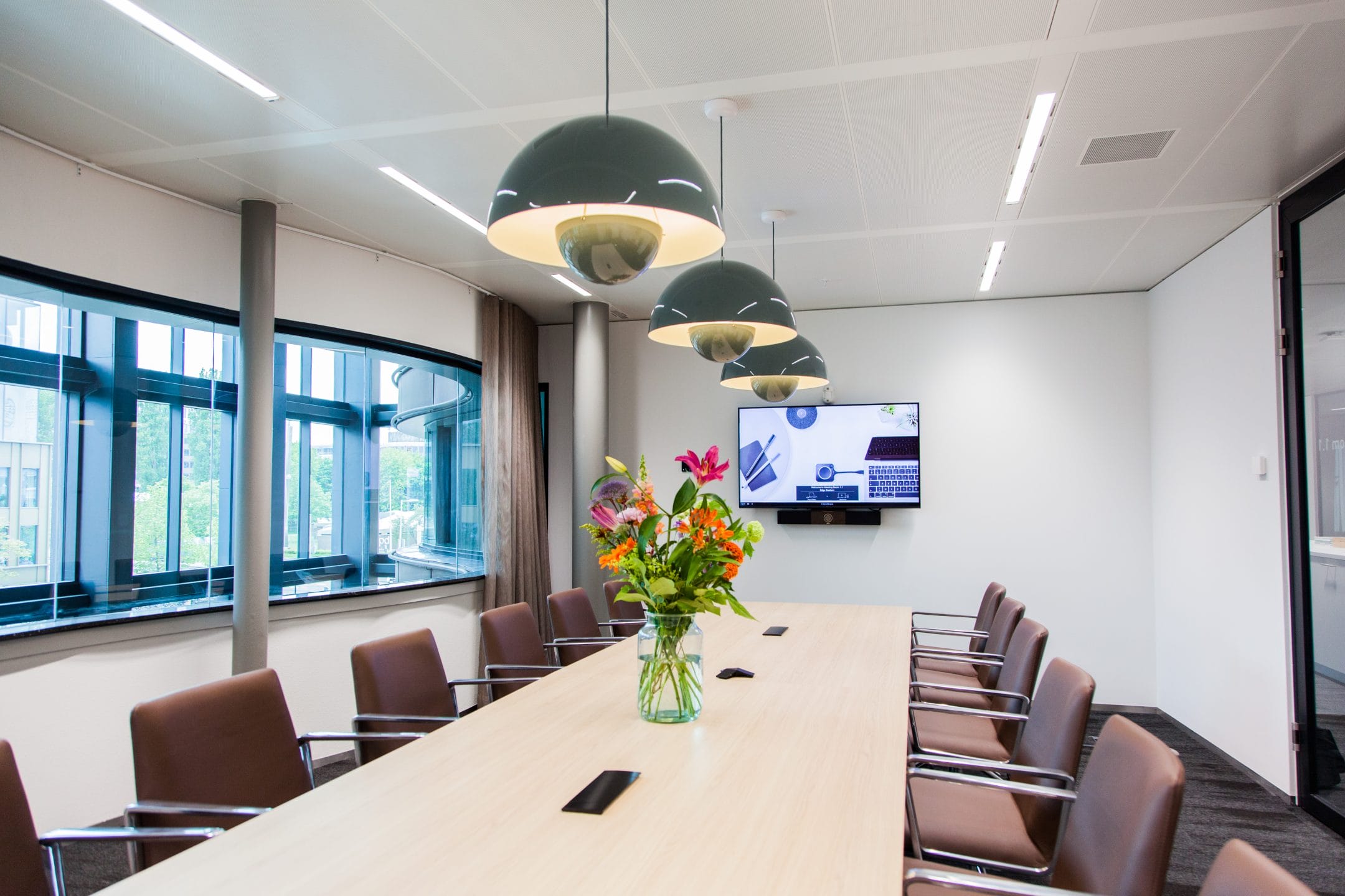 A modern conference room with a long table, surrounded by brown chairs. There's a vibrant flower arrangement in the center. Overhead, unique green pendant lights hang. A large monitor displays a presentation on An image from EDGE Workspaces.