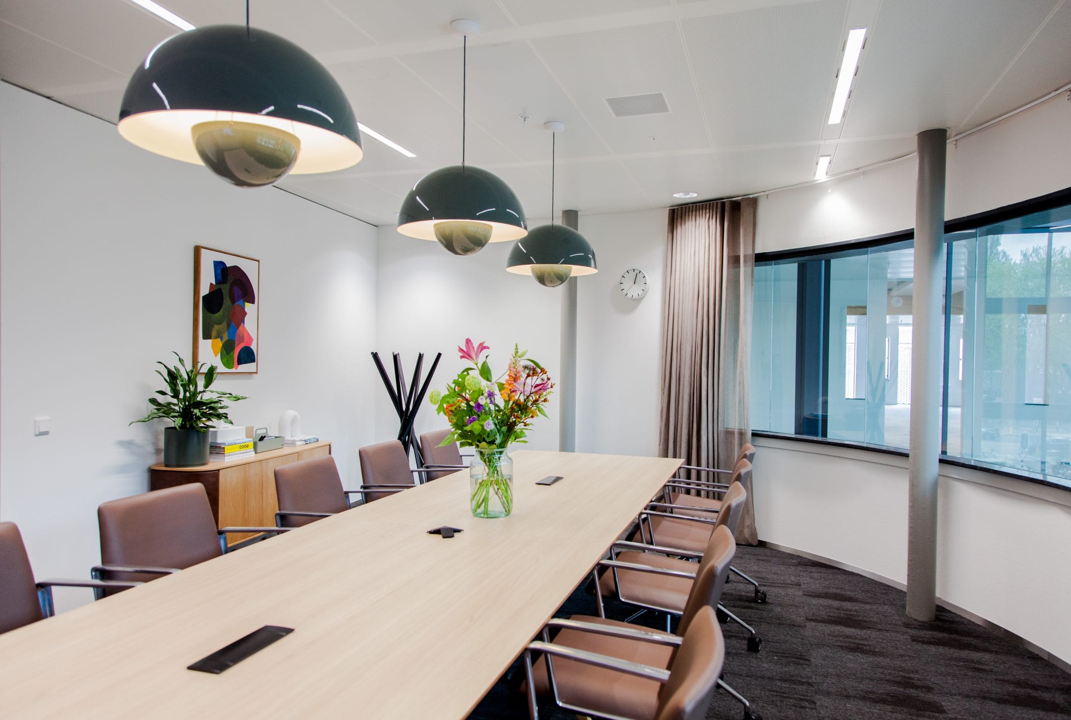 Ein moderner Konferenzraum mit einem langen Tisch mit Stühlen, drei großen Hängelampen darüber, einem farbenfrohen abstrakten Gemälde an einer Wand und einer Vase mit frischen Blumen auf dem Tisch. Große Fenster sorgen für natürliches Licht. Ein Bild von EDGE Workspaces.