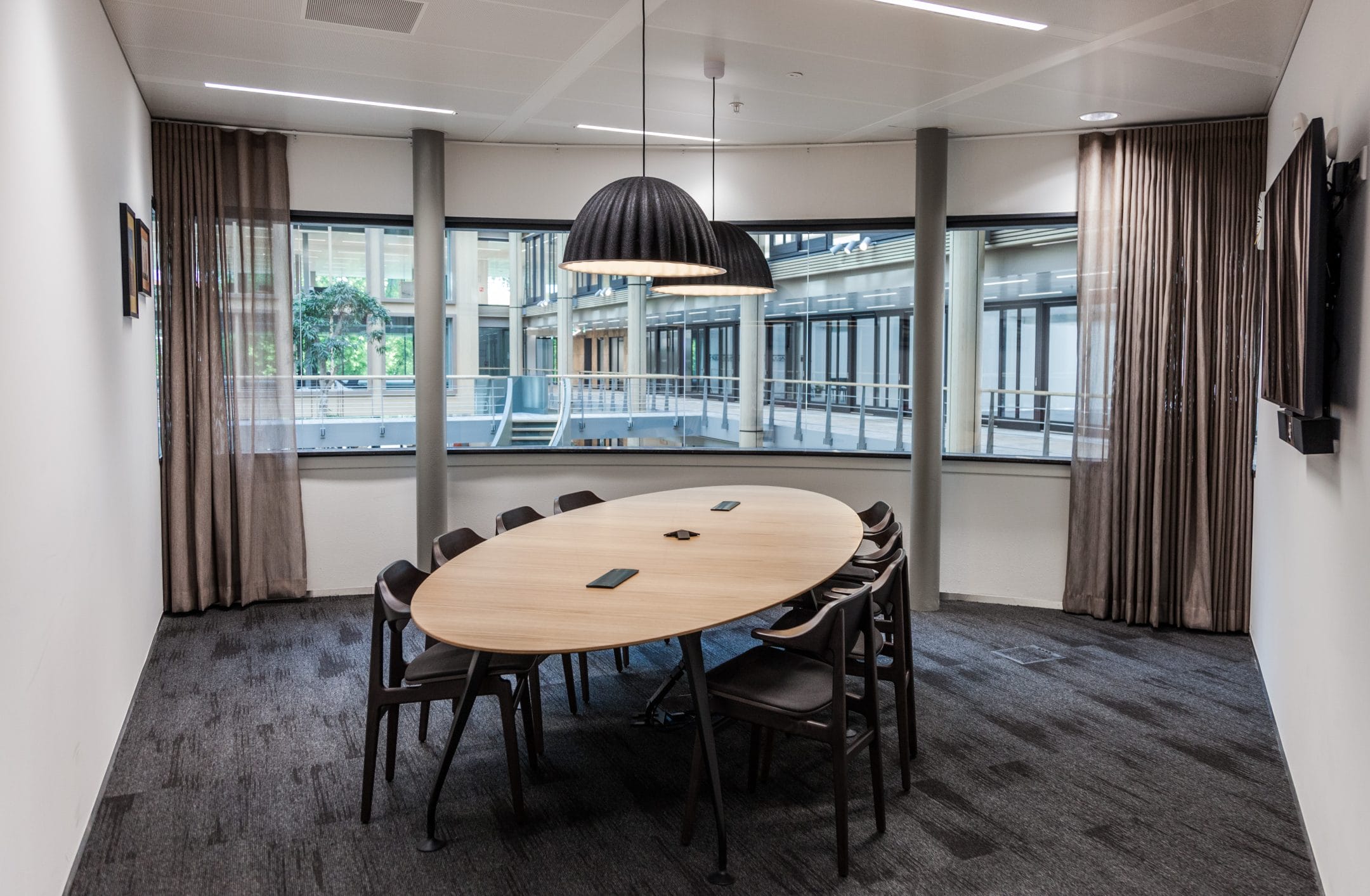 Modern coworking space featuring a large oval wooden table with chairs, a unique black pendant lamp, floor-to-ceiling windows with city view, curtains, and carpeted floor. An image from EDGE Workspaces.