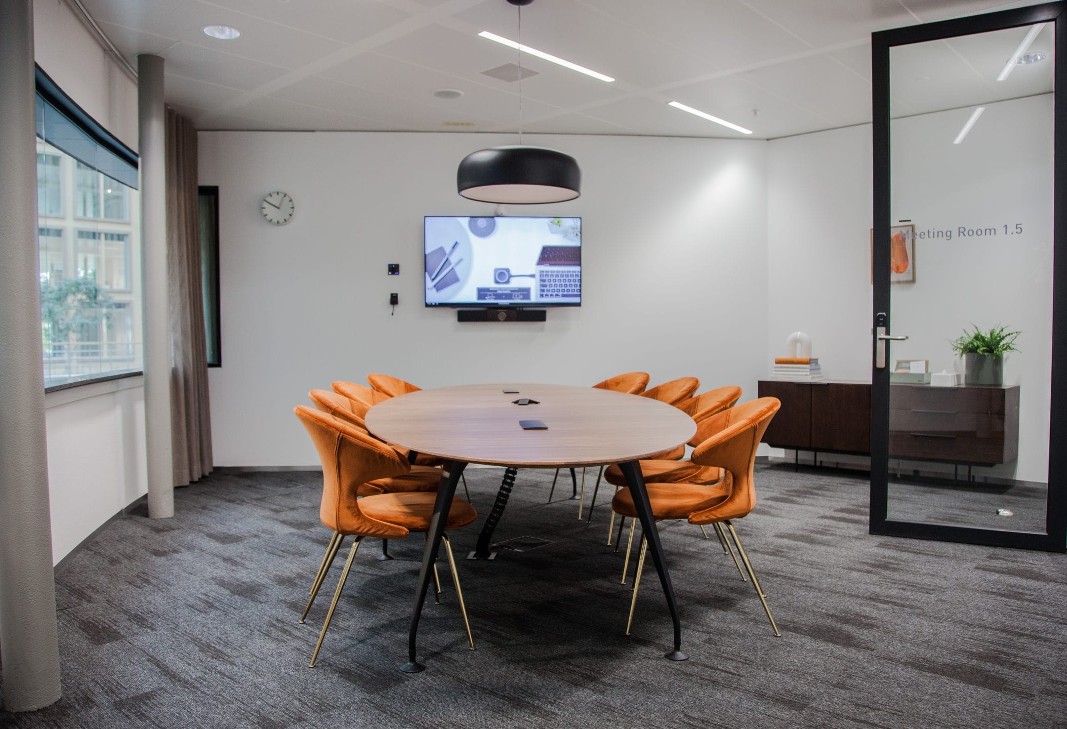 Modern office meeting room with an oval table surrounded by orange chairs, a TV on the wall displaying a presentation, a circular ceiling light, and a glass door labeled "coworking space 1. An image from EDGE Workspaces.