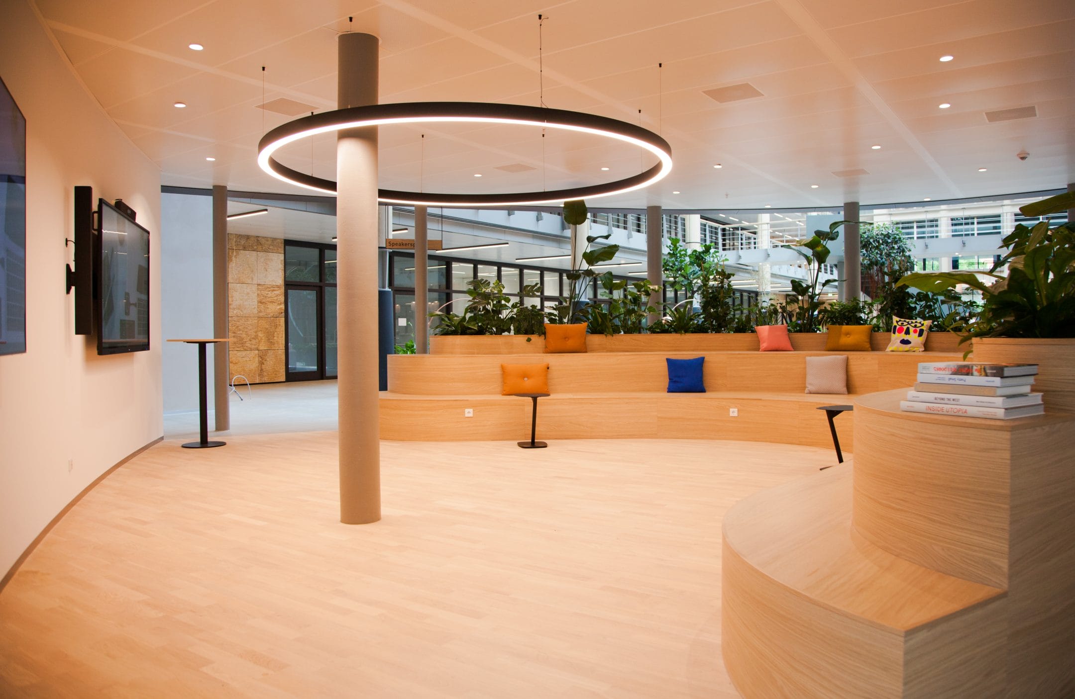 A modern office lobby with a wooden circular seating area, a large ring light overhead, and plants in colorful pots, with books on a table, and large windows in the background offering access to flexible workspace An image from EDGE Workspaces.