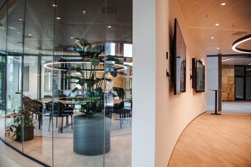 Moderne Büroloby mit gebogenen Glaswänden, die einen Sitzbereich mit Grünpflanzen umschließen, und einem Flur mit gerahmten Kunstwerken an den Wänden, die ein sauberes und zeitgemäßes Design widerspiegeln, ideal für innovative Coworking-Umgebungen. Ein Bild von EDGE Workspaces.