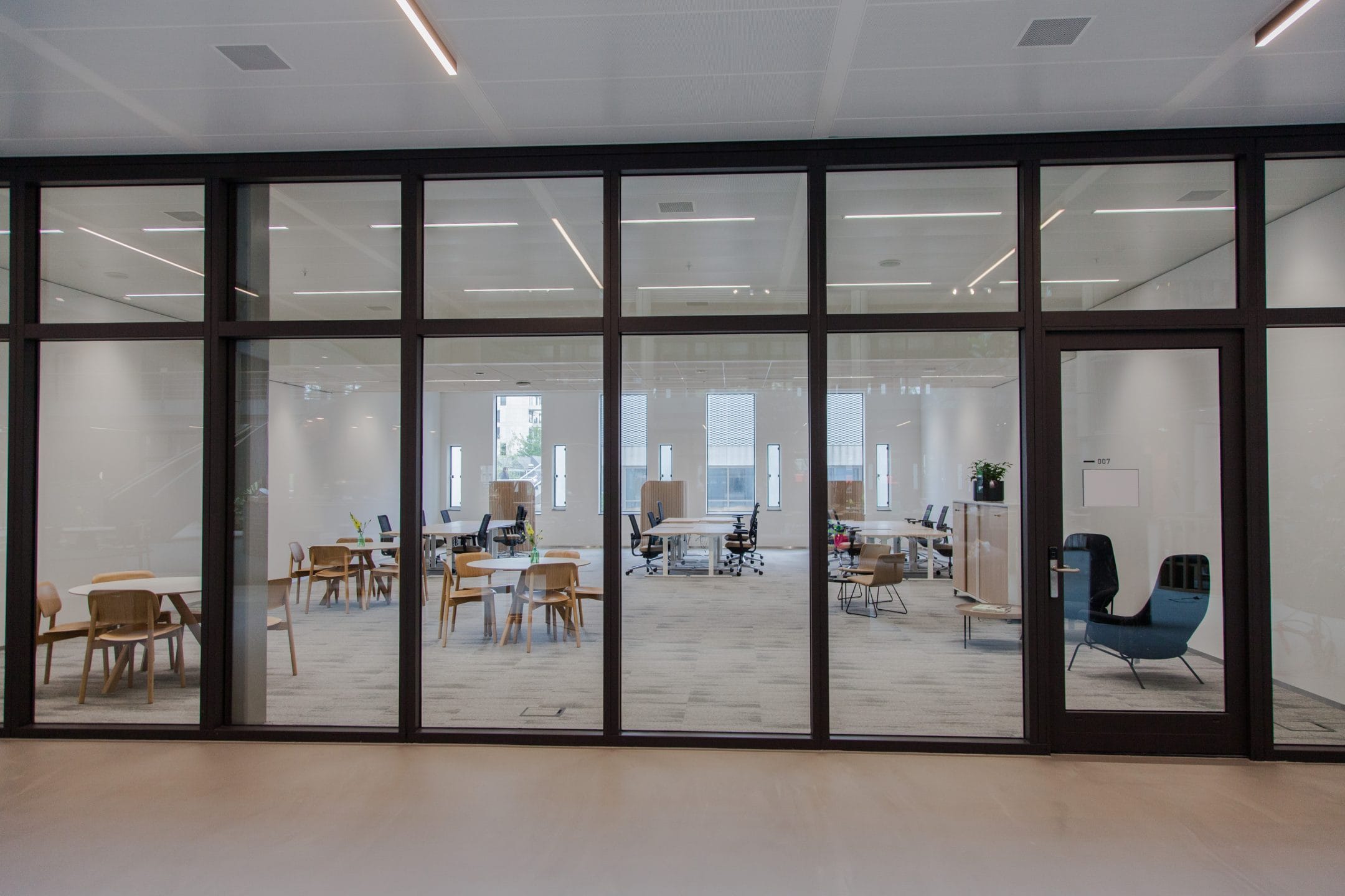A modern office interior seen through large glass windows features scattered seating arrangements with tables, chairs, and a few sofas. Bright natural light illuminates the space, highlighting a clean, minimalistic design tailored for An image from EDGE Workspaces.