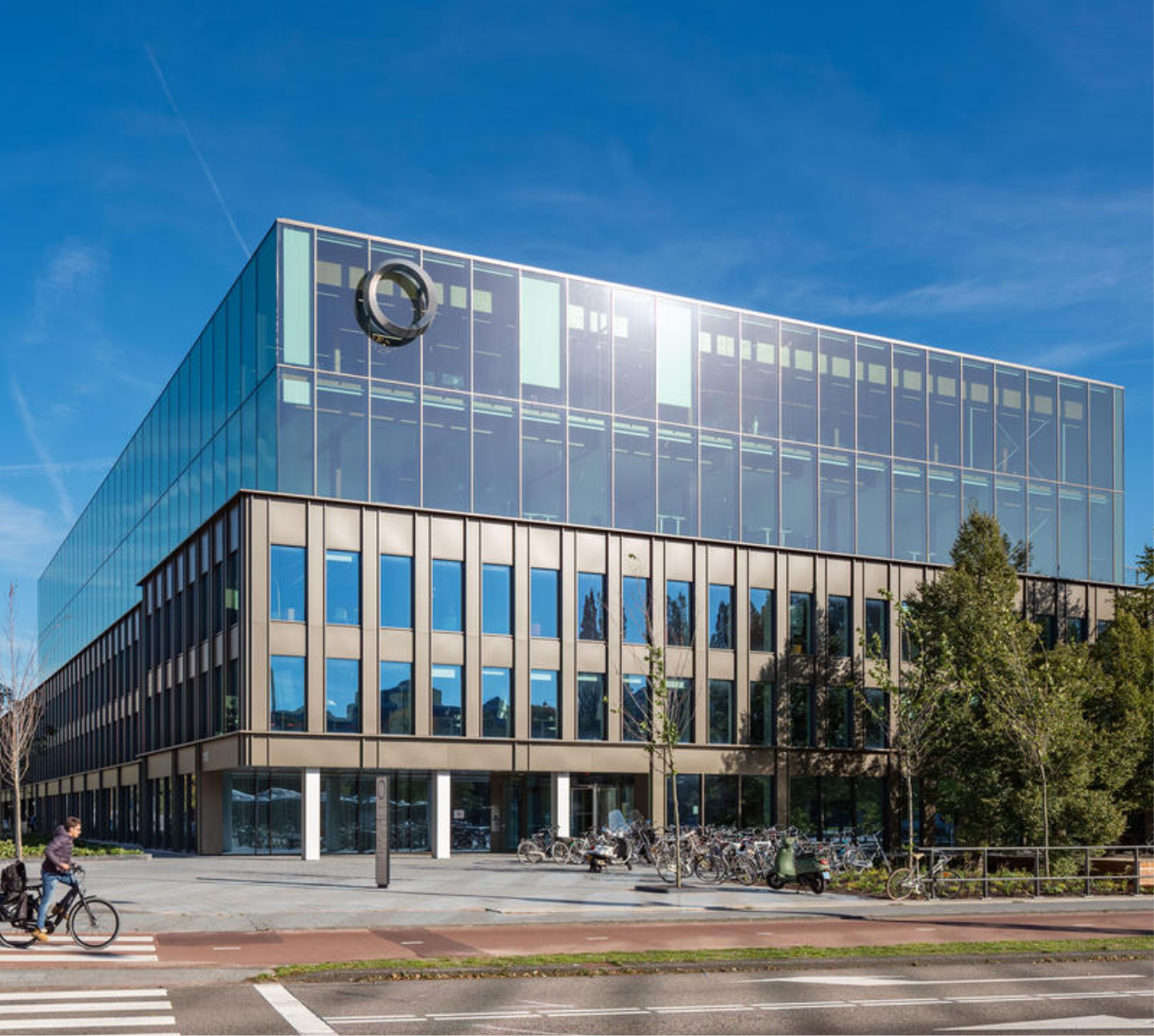 Ein modernes, reflektierendes Glasgebäude mit einer verspiegelten Fassade, die einen klaren blauen Himmel und Bäume zeigt. Die Struktur ist als innovativer Coworking Space mit einem geometrischen Design mit horizontalen und vertikalen Linien konzipiert. Ein Radfahrer Ein Bild von EDGE Workspaces.