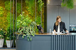 A professional woman reading a book at a modern reception desk in an innovative coworking environment, surrounded by lush greenery and hanging plants. The area features stylish gray and green decor with a tranquil, natural An image from EDGE Workspaces.