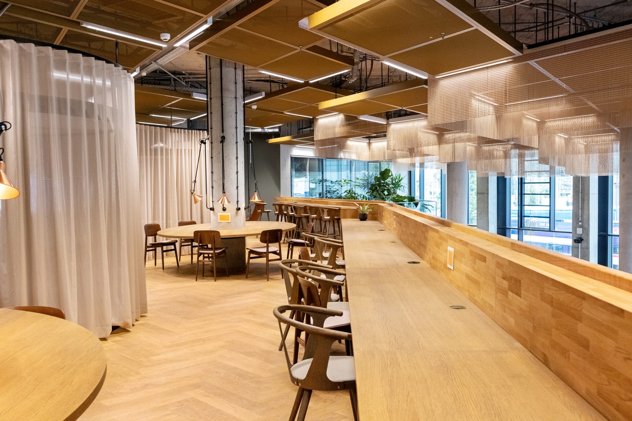 A modern office break room featuring wooden tables and chairs, with a long bar area on the right. The room has many pendant lights and green plants by large windows, complemented by soft, white curtains An image from EDGE Workspaces.