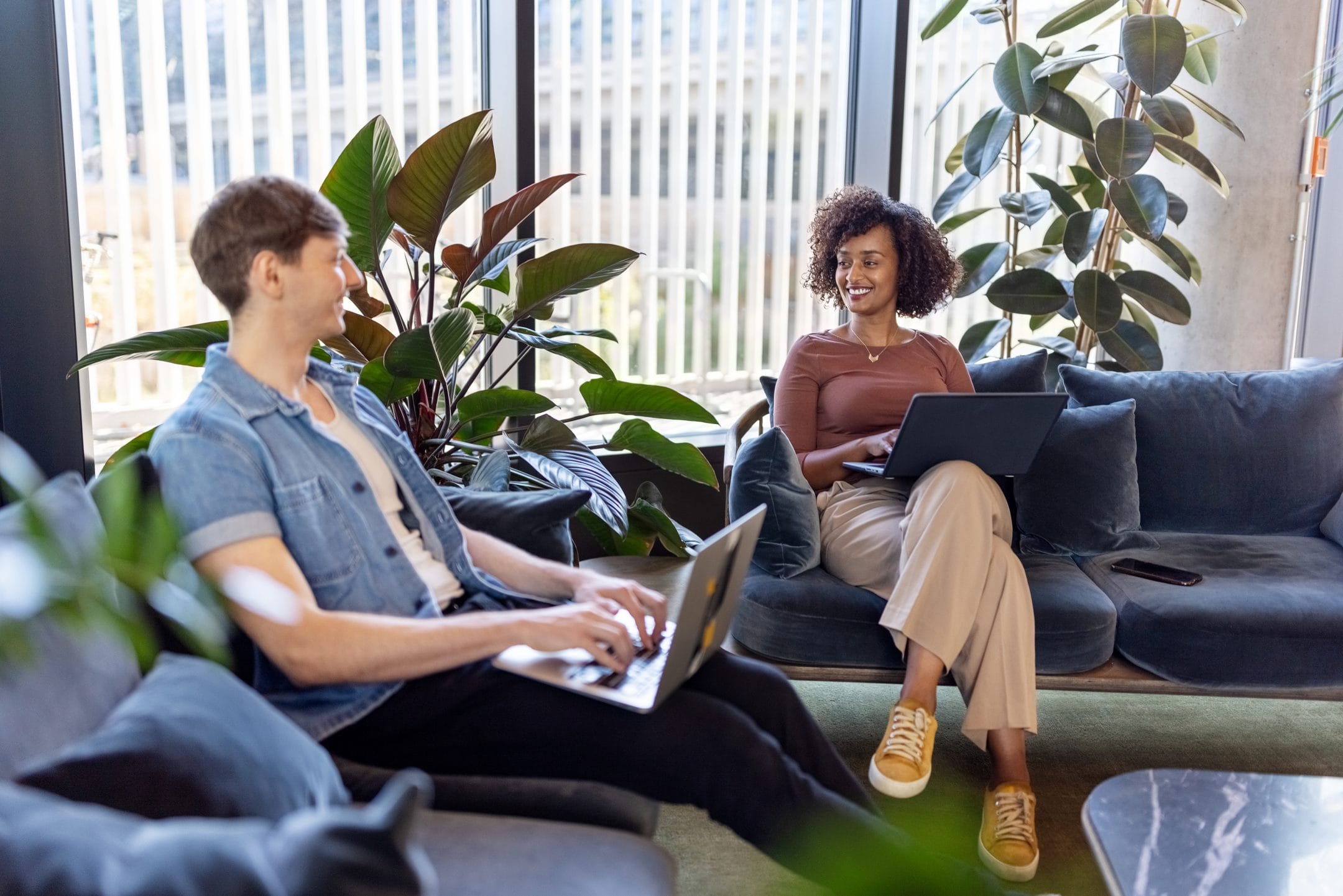 Two people, a man and a woman, are sitting in a modern coworking lounge area with laptops. The woman, seated on a couch, smiles at the man across from her who sits on a An image from EDGE Workspaces.