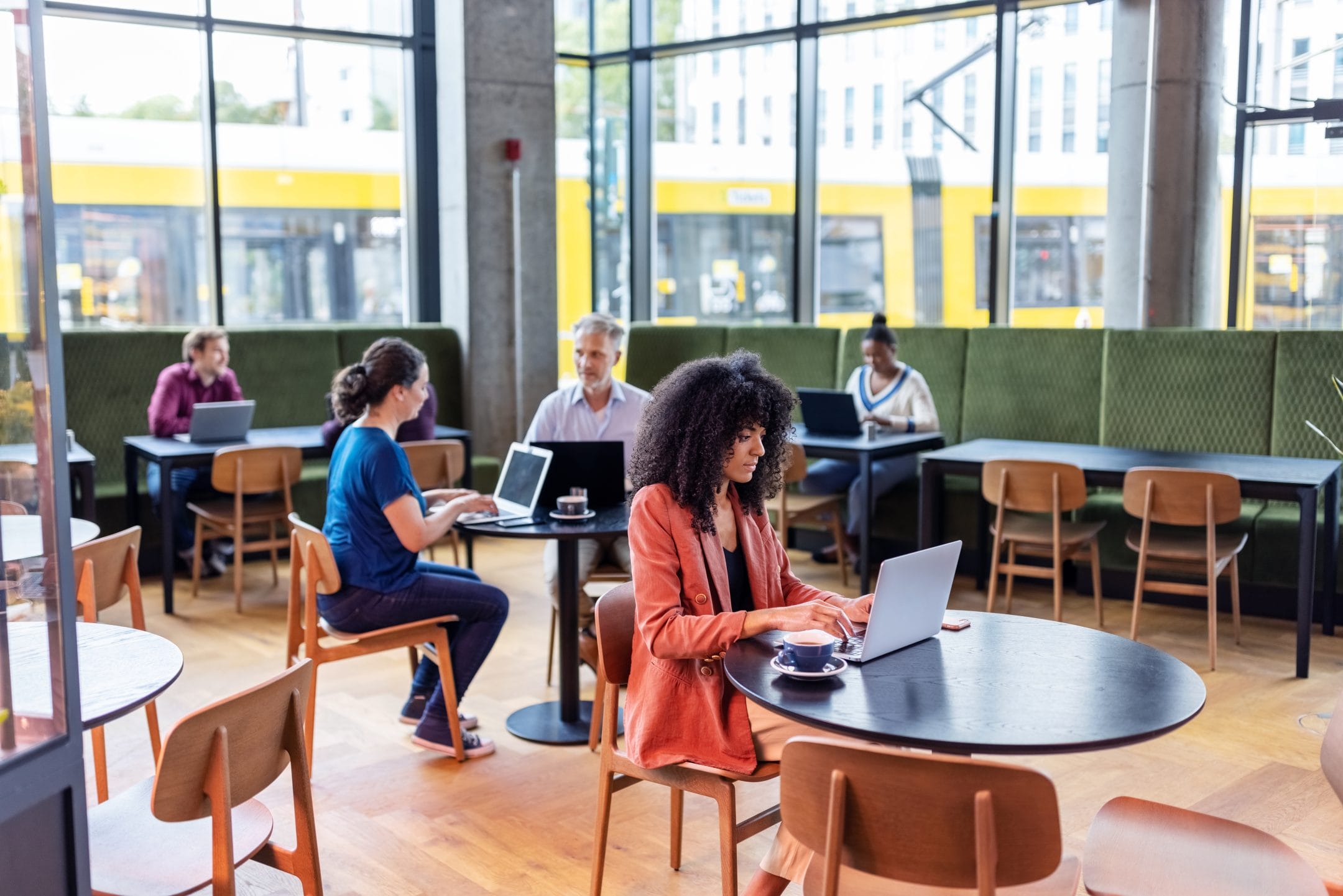 A young woman with curly hair works on a laptop in a modern café equipped with dedicated workspaces, sitting at a table with a cup of coffee. Behind her, various people are also working on laptops An image from EDGE Workspaces.