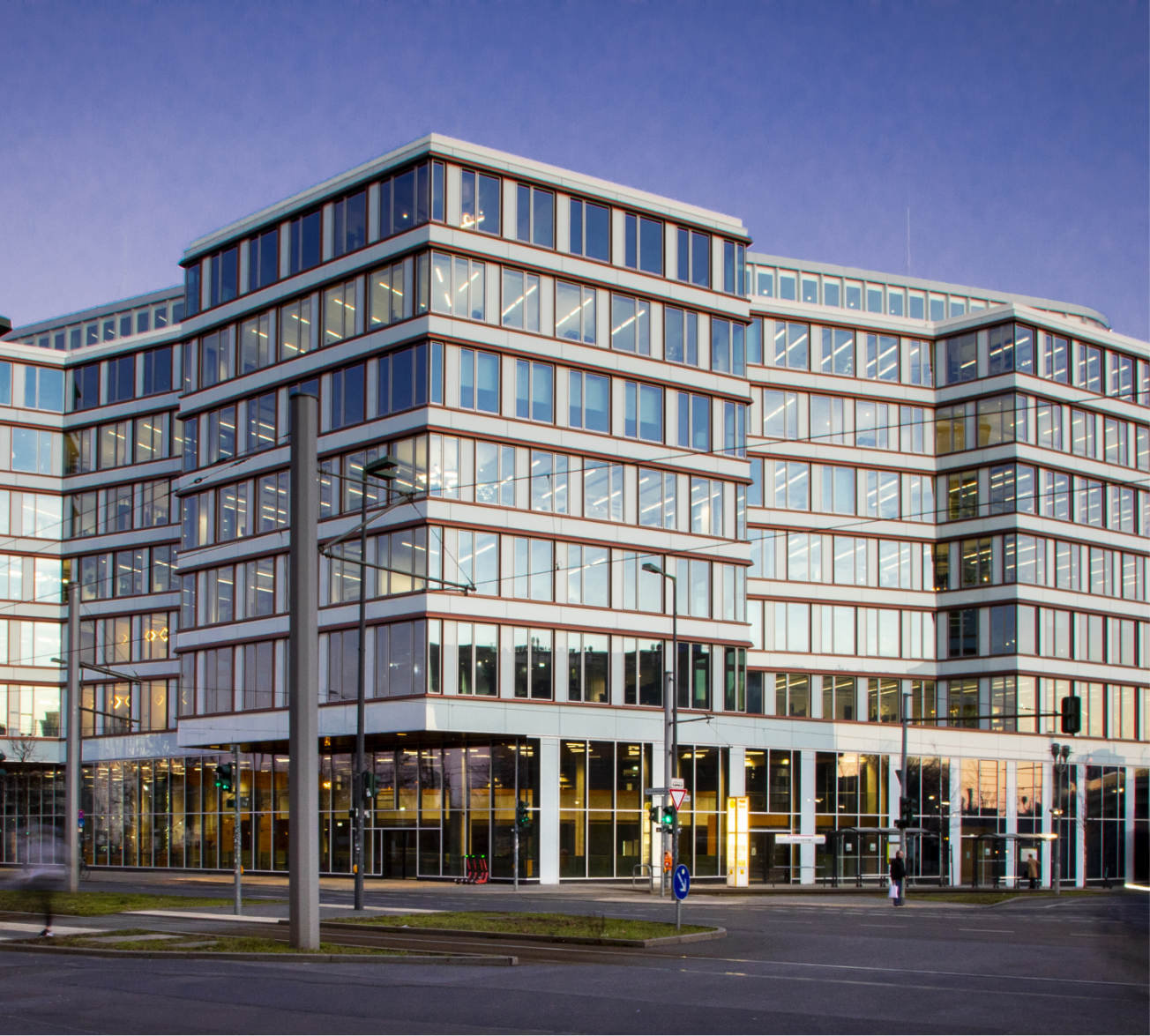 Modernes Bürogebäude mit weitläufiger Glasfassade und mehrstufiger Struktur, aufgenommen in der Abenddämmerung. Das Gebäude bietet nachhaltige Bürolösungen mit einem rechteckigen Design und markanten weißen Rahmen, an einer Straßenecke unter klarem Himmel gelegen. Ein Bild von EDGE Workspaces.
