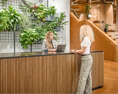 Zwei Frauen mit lockigem blonden Haar an einem modernen Büroempfang. Eine sitzt hinter einem Schreibtisch mit einem Laptop und Pflanzen darüber, die andere steht am Schreibtisch. Der Bereich verfügt über hölzerne Designelemente und ist Teil eines Bildes von EDGE Workspaces.