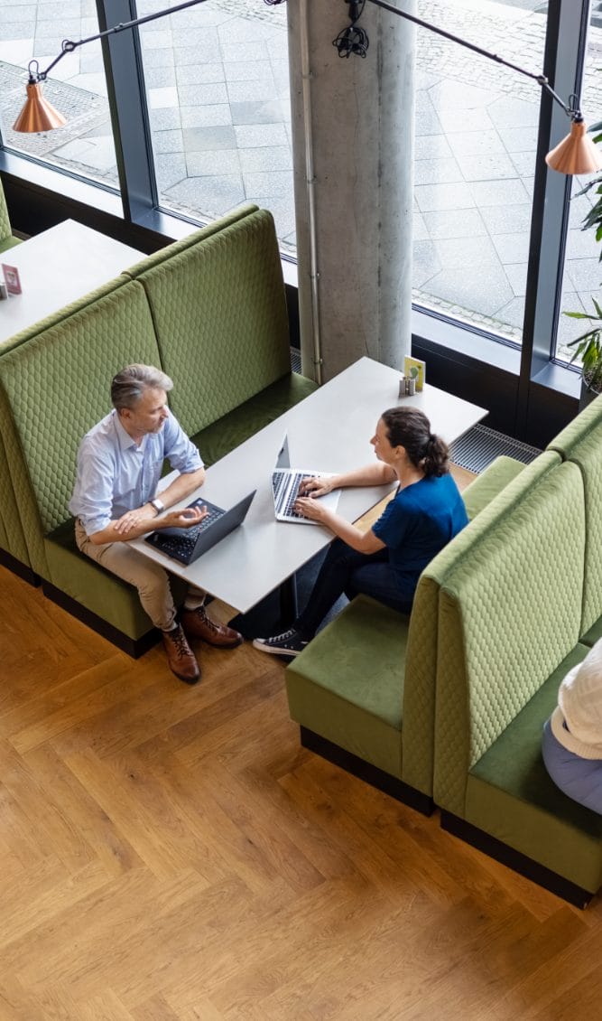 Two people, a man and a woman, are seated across from each other at a booth with laptops in a bright, modern coworking space with large windows and hanging lights. They appear to be engaging An image from EDGE Workspaces.