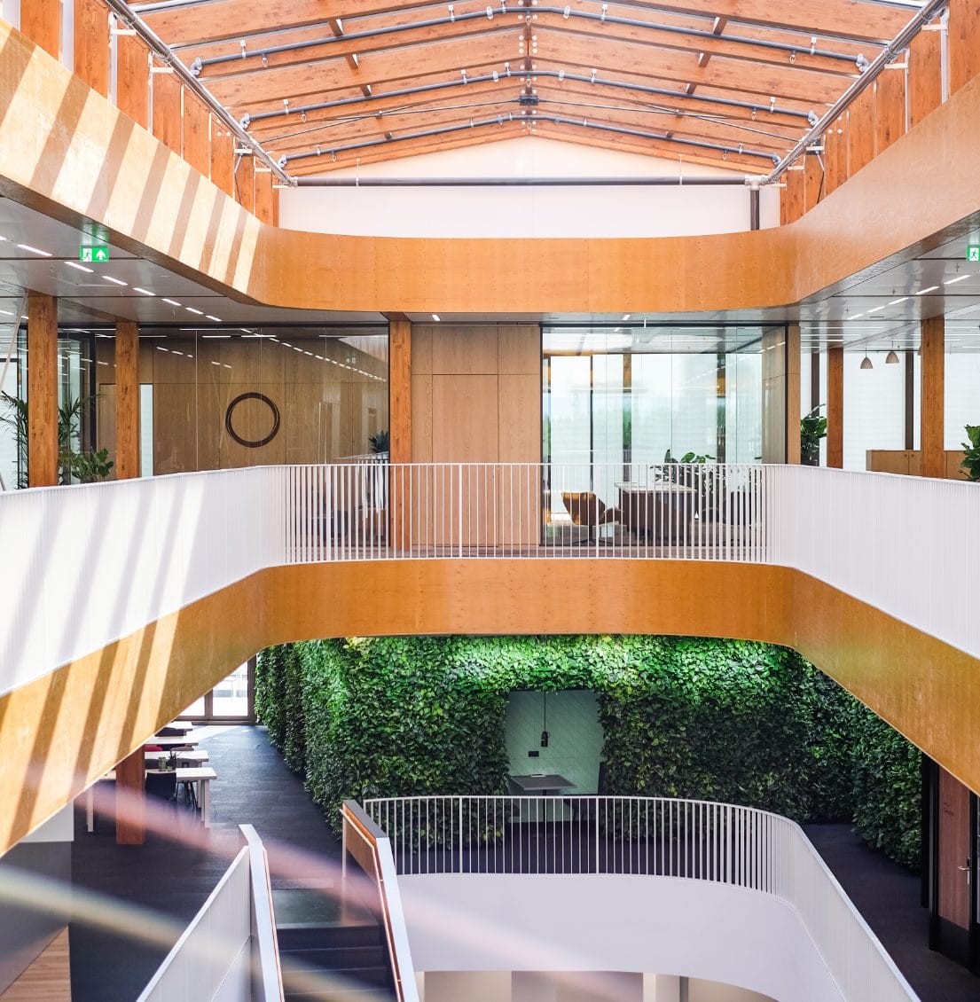 Modernes Bürogebäude-Interieur mit geschwungenen weißen Gehwegen, Holzvertäfelung und einer üppigen grünen Pflanzenwand. Das Design zeichnet sich durch viel Tageslicht, minimalistisches Dekor und innovative Coworking-Umgebungen aus. Ein Bild von EDGE Workspaces.