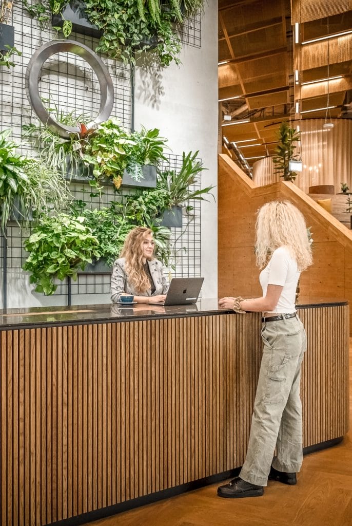 A woman with curly blonde hair standing at a wooden reception desk in an innovative coworking environment, speaking to another woman with curly blonde hair using a laptop. The background features a green wall with plants and An image from EDGE Workspaces.