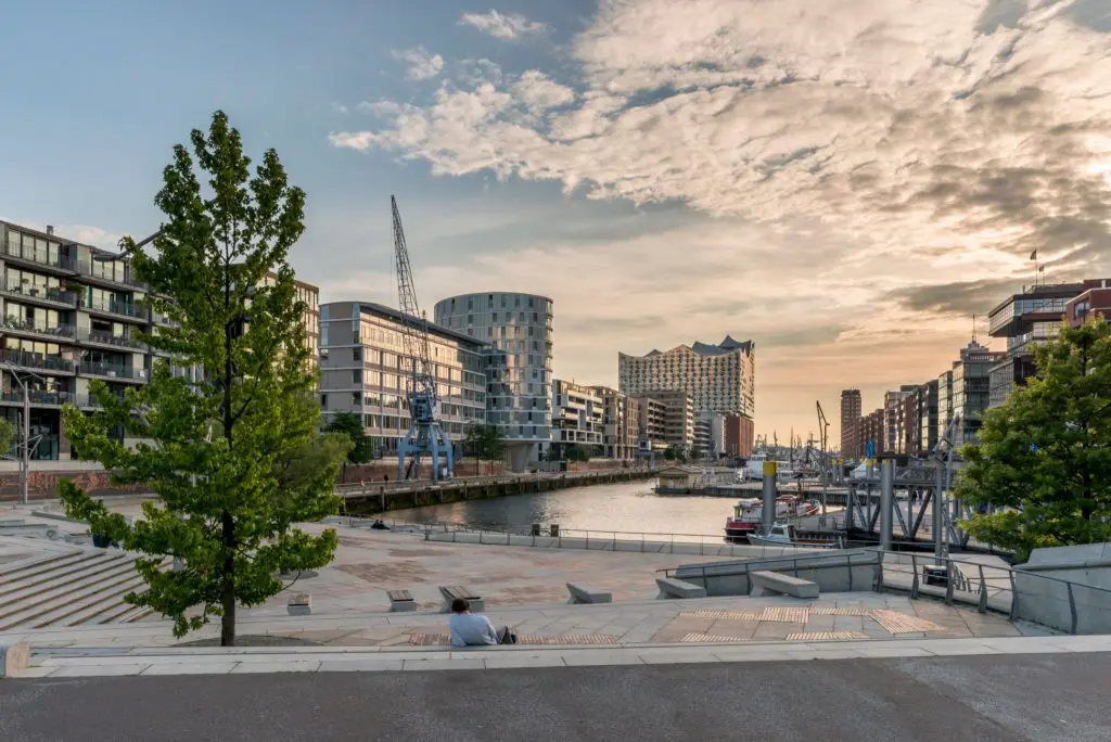 A waterfront promenade at dusk with modern buildings and a calm channel of water. Memberships are available for the event spaces nestled among the buildings, where a few people are sitting and one walks along the An image from EDGE Workspaces.