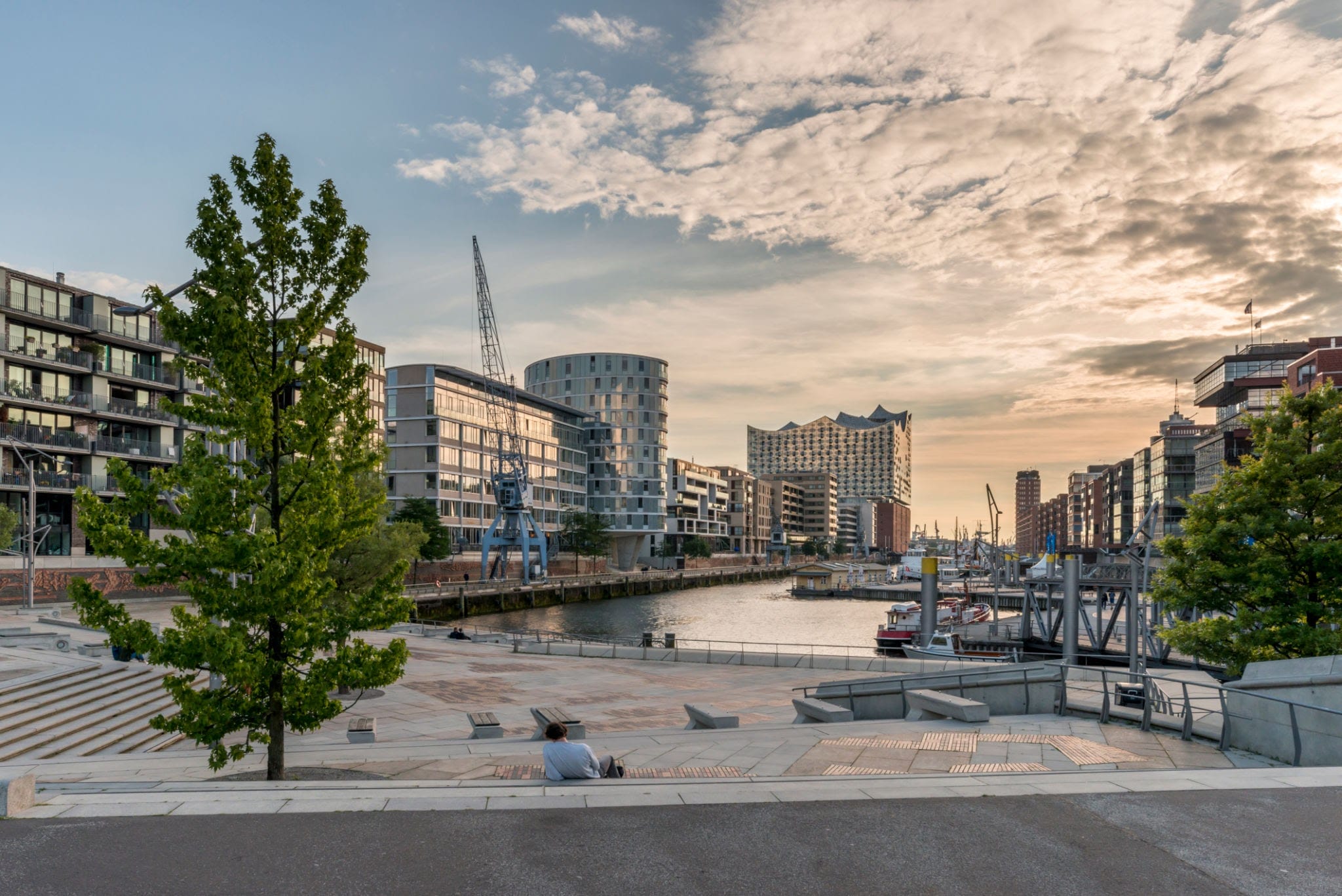 A waterfront promenade at dusk with modern buildings and a calm channel of water. Memberships are available for the event spaces nestled among the buildings, where a few people are sitting and one walks along the An image from EDGE Workspaces.