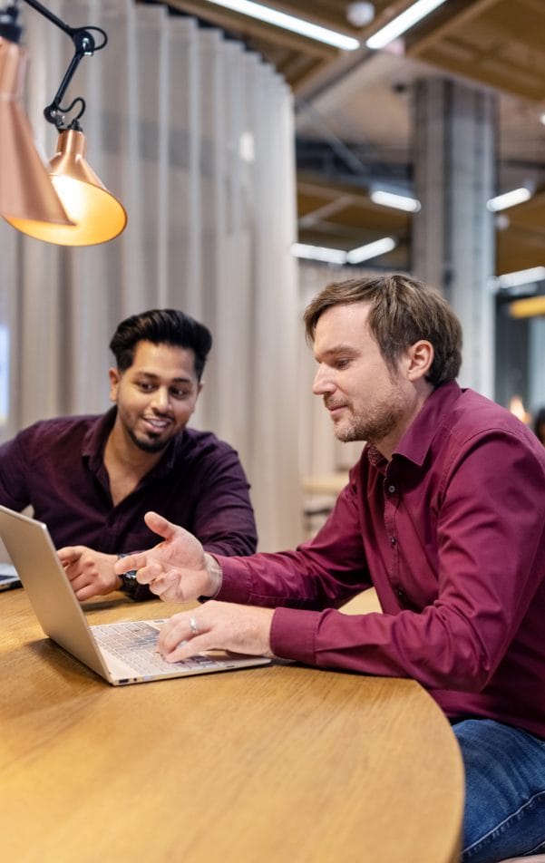 Zwei Männer, einer mit dunklen und einer mit hellen Haaren, sitzen an einem Holztisch in einem gut beleuchteten Private Office. Sie sind in ein Gespräch vertieft, wobei ein Laptop vor ihnen geöffnet ist. Ein Bild von EDGE Workspaces.