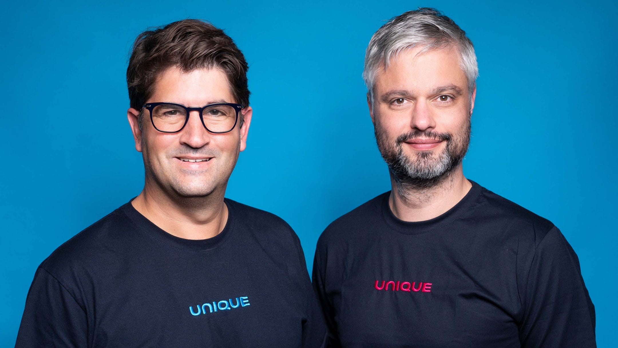 Two smiling males in black t-shirts labeled "unique" standing side by side against a vibrant blue backdrop in a coworking space. The man on the left wears glasses and has dark, wavy hair An image from EDGE Workspaces.